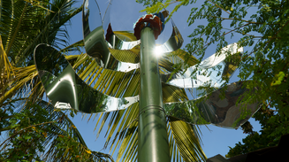 Skulpturel, palmelignende metalinstallation på høj mast, der reflekterer sollys mellem grønne træer og blå himmel.