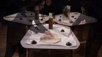 Kunstinstallation med hænder, der interagerer med objekter på et geometrisk bord i dæmpet lys.