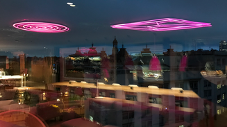  Neonlys i loftet spejler sig i glas med udsigt over byens tage i skumringen.