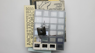 A stack of archival material rests on a light surface, topped with a clear sheet of photo slides arranged in a grid. A small black slide viewer rests on top, and sticky notes stick out from the pages. 