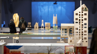 Arkitekturmodeller og tegninger fra en udstilling med besøgende i baggrunden.