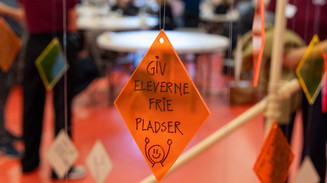 Orange skilt med teksten "Giv eleverne frie pladser" hænger foran en sløret workshop.