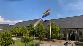 Et regnbueflag på flagstang vajer over solbeskinnet campus
