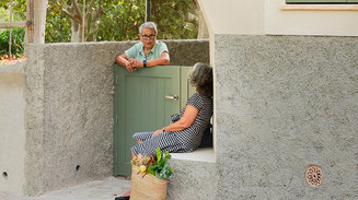 Two women chat over a low garden gate outside a house, with a shopping basket on the ground.