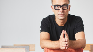 Man in black glasses leans over the back of a wooden chair.