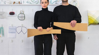 Two people in black hold wooden pieces in a design studio as a dog looks on.