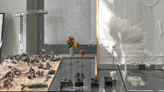 Installation med skråt modelterræn, gennemsigtige flader, metalstativer og organiske skulpturer i et lyst udstillingsrum.