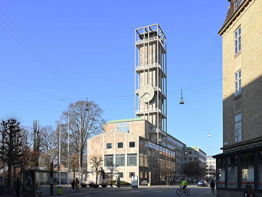 Høj bygning med urtårn ved en bygade under blå himmel.