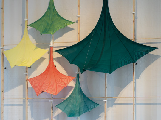 Five colorful, funnel-shaped textile installations hang from a wooden frame against a white wall.