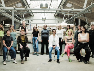 Group portrait of people seated and standing in a bright workshop with shelves, tools, and industrial lamps in the background