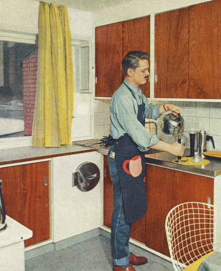 Mand hælder kaffe i en kop i et køkken med træskabe og gule gardiner.