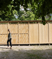 Kvinde går forbi en trækonstruktion med glatte og flettede paneler under grønne træer.