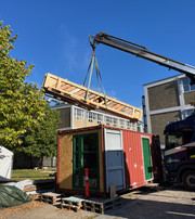 En kran løfter en lang trægitterkonstruktion over en rød container ved en industribygning.