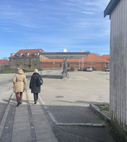 To personer går på fortovet forbi en åben plads med en ladestander og røde murstensbygninger.