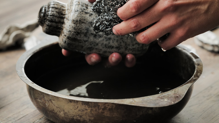 Hands cleaning a ceramic vessel over a bowl of water.