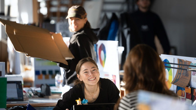 Personer samarbejder og taler sammen i et lyst atelier med farvecirkler og designskitser.