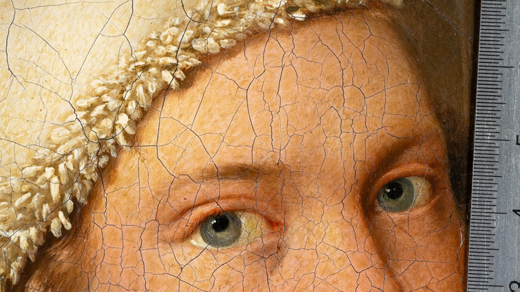 Detail of a cracked painting showing two blue-grey eyes and a ruler along the right edge.