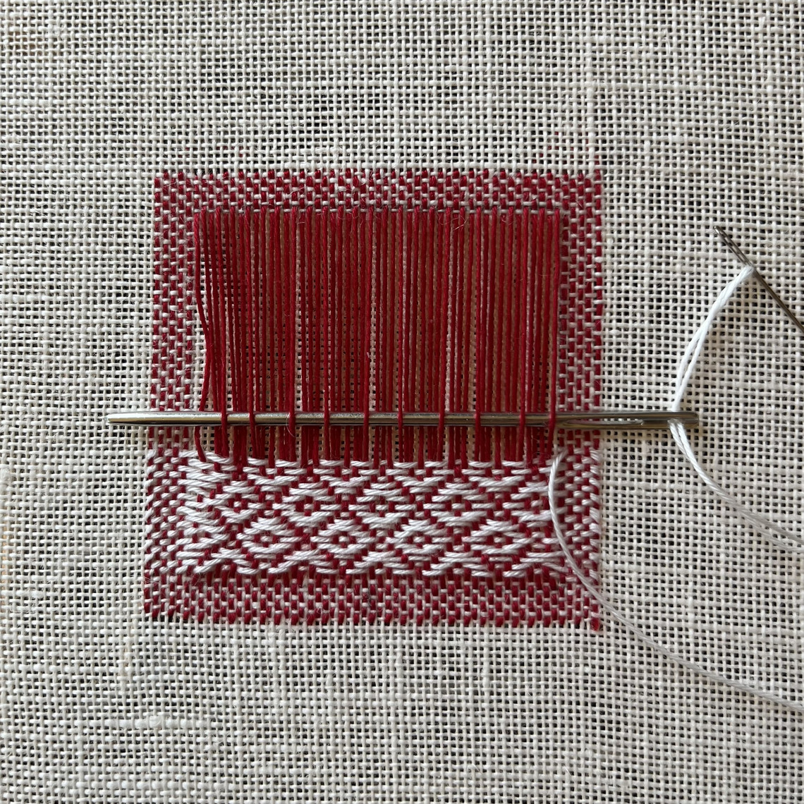 Image displaying a half-finished textile darn on an off-white linen. The darn is sewn with red yarn vertically, and white yarn horizontally and in the woven structure called ‘diamond twill’. 