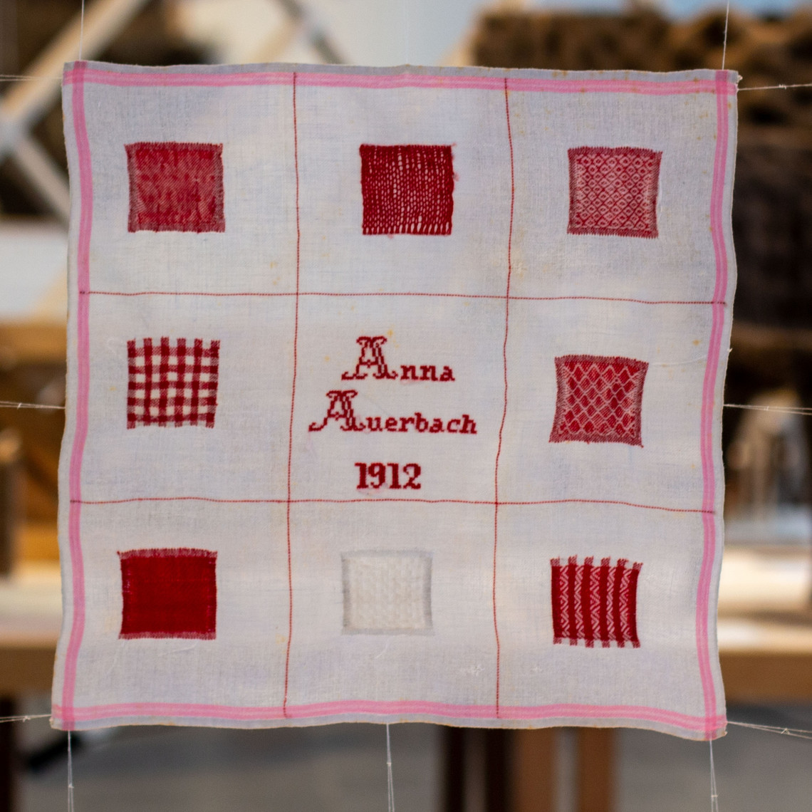 The image displays a darning sampler. The sampler is an off-white square piece of linen on which eight different darns in red and white are sewn. The darns are organized in a three-by-three grid. In the middle of the grid, the text “Anna Auerbach 1912” is sewn with cross-stitches.