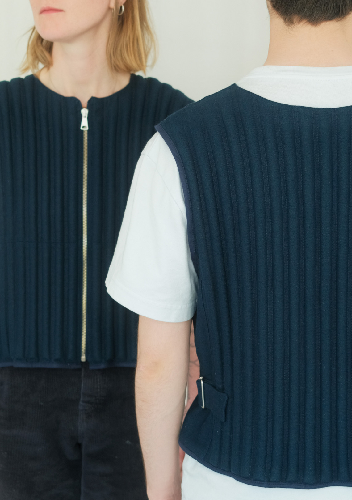 Close-up of two people wearing dark pleated vests, showing front and back details.