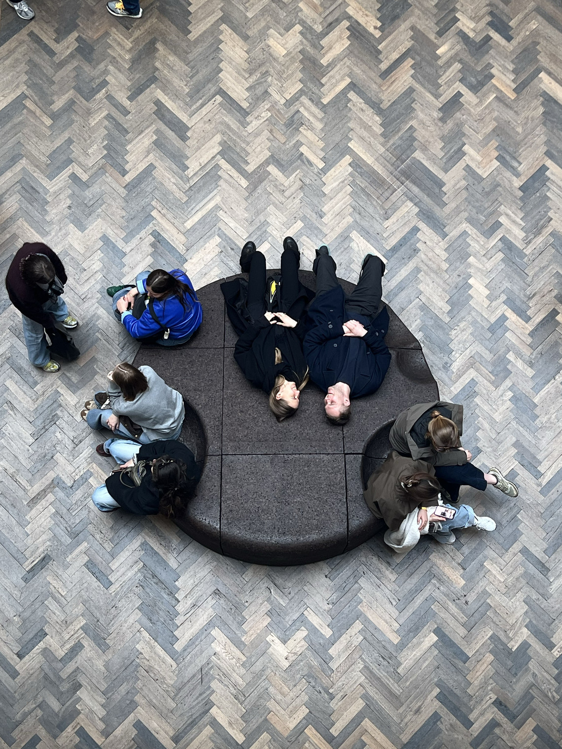 People sitting and lying on a circular bench surrounded by herringbone-patterned flooring.