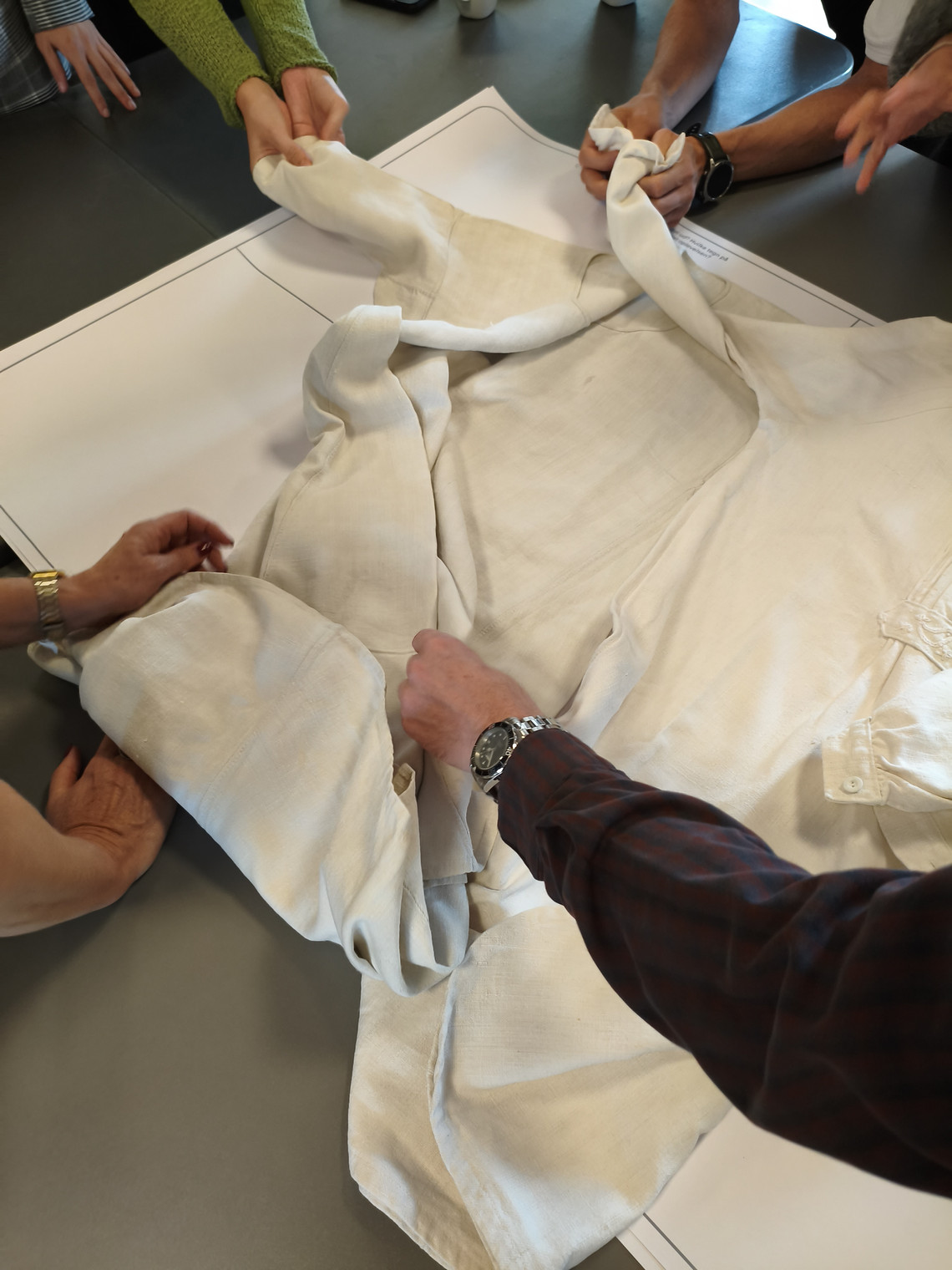 A white cloths lays on a table and many hands are holding it
