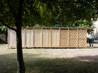 Long timber pavilion with varied facade cladding beneath large trees in a courtyard.