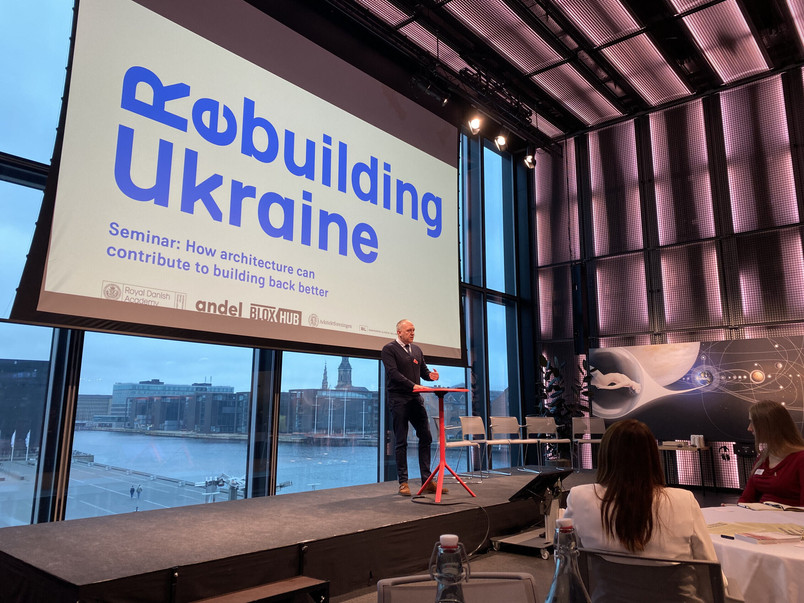 Oplægsholder står på en scene foran et stort lærred med teksten “Rebuilding Ukraine” under et seminar.