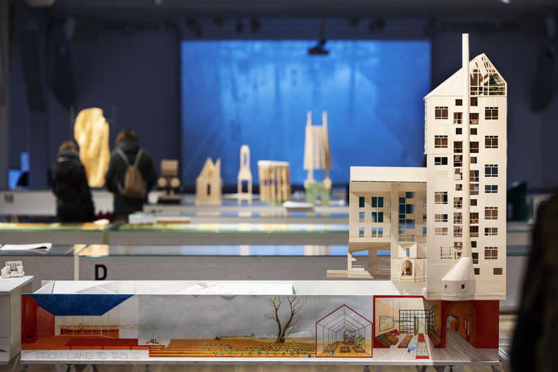 Arkitekturmodeller og tegninger fra en udstilling med besøgende i baggrunden.