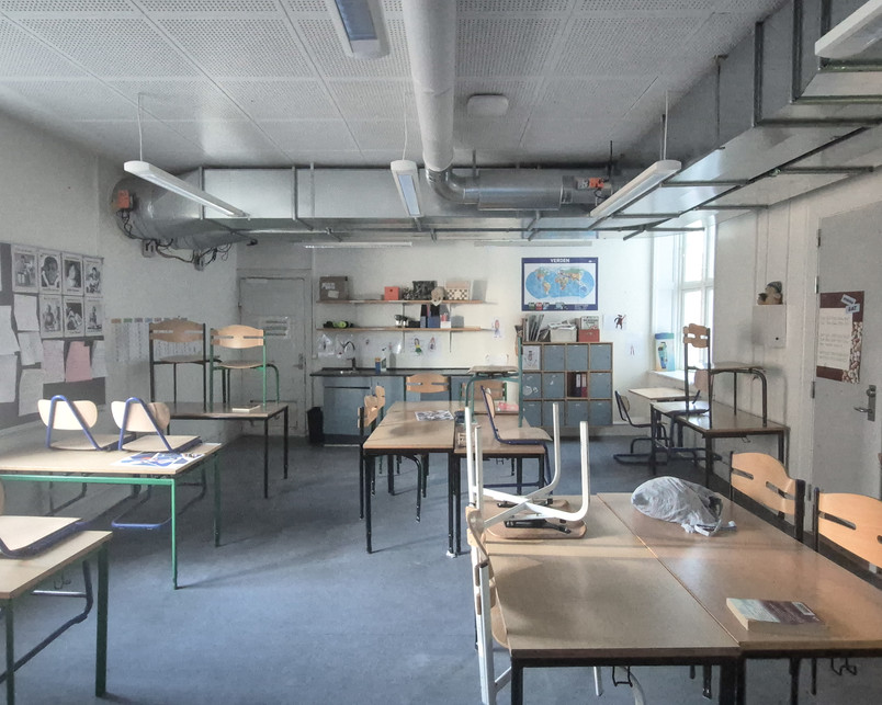 Photo of a classroom: Inside one of the classroom, the smooth walls and matte gray floor is repeated. The ceiling is crossed by very large exposed pipes. Located between two disconnected hallways, the room is left as a passage. Desks are spaced out with some chairs flipped on top. Shelves, posters, and a world map line the back wall.