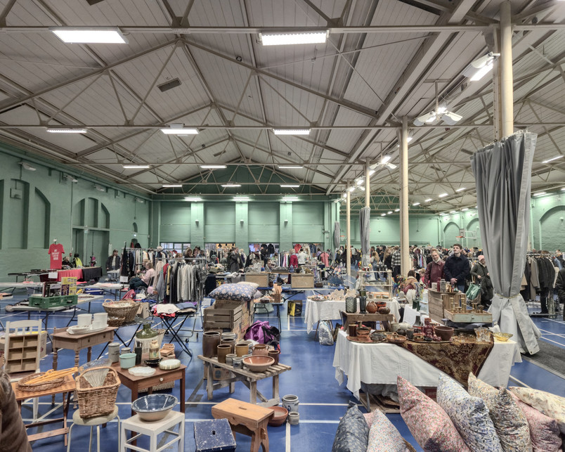Photo inside Remisen sportshall. Inside Remisen, the big sports hall is lined up with long rows of tables for a sunday flea market. The windows are permanently blinded and the indoor lighting from above feels artificial.
