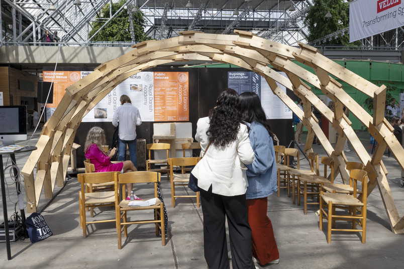 Wooden arch structure with chairs underneath, surrounded by exhibition displays and visitors.