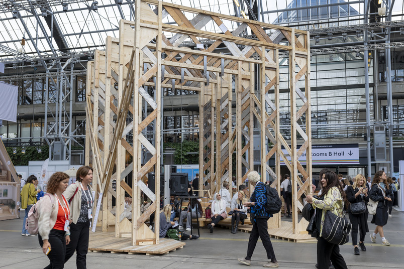 Tall timber pavilion structure with people walking and sitting inside a busy exhibition hall.