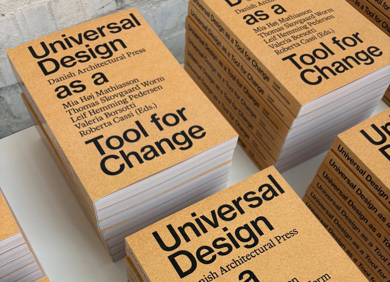 The picture shows piles of physical copies of the Anthology. The cover of the Anthology has a light brown background with a black and bold text: Universal Design as a Tool for Change