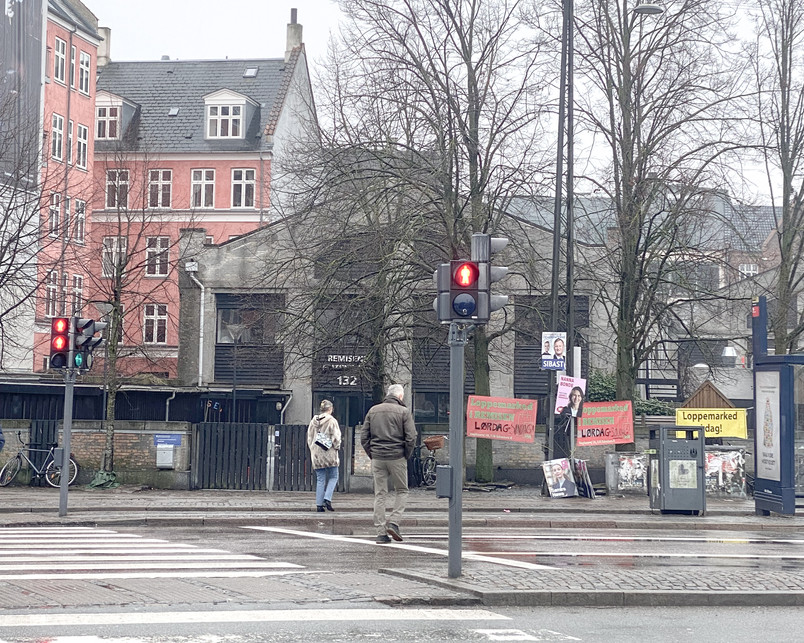 Photo of Remisen seen from the metrostop Trianglen. Standing with our back to Trianglen Metro facing a busy traffic junction on Blegdamsvej. Across the pedestrian crossing, Remisen faces us with a closed off facade.  Surrounded by the rythmic noises of car breaks whining, the traffic light beeping and engines accelerating. The wind is coming from all sides in this open space.