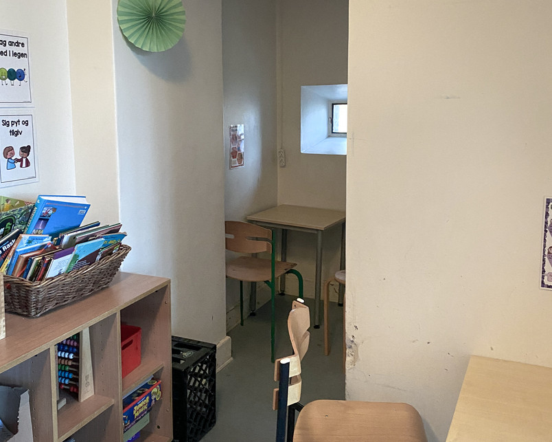 Photo of a small reading nook in the corner of a classroom: In the corner of a classroom there is a small isolated seating area. A table and chair is facing a wall with a tiny window above eye height. The space is dimly lit and it feels dark and neglected. It looks more like a punishment than a space to gather your thoughts.
