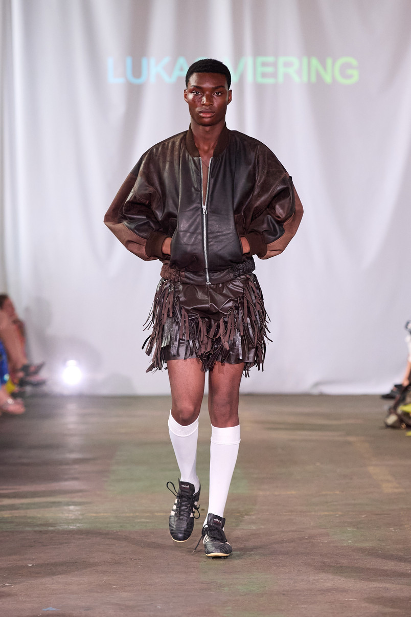 Model på catwalk iført mørk læderjakke, frynset nederdel, hvide knæstrømper og sorte sneakers.