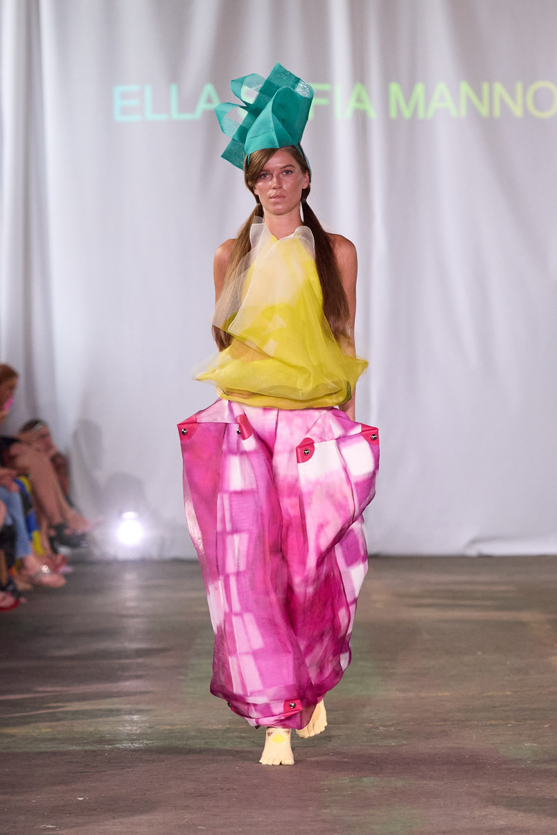 Model på catwalk iført transparent gul top, voluminøse pink bukser og en stor turkis hovedbeklædning.