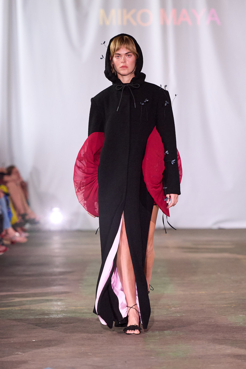 Model på catwalk iført lang sort kjole med hætte og røde, skulpturelle sidepaneler.