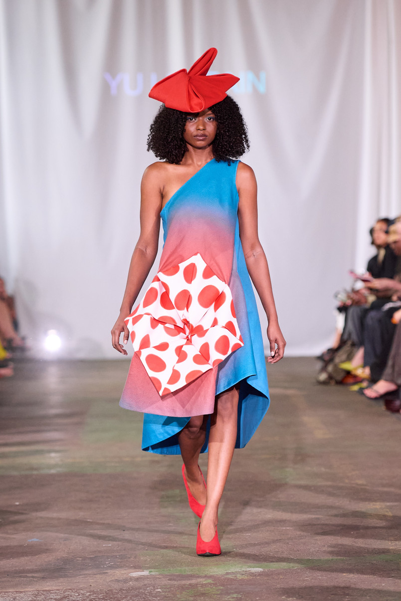 Model på catwalk iført asymmetrisk blå-rosa kjole med rødprikket skulpturelt panel og stor rød hat.