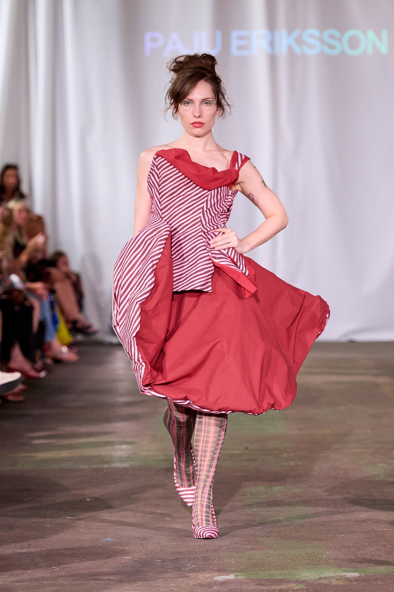 Model på catwalk iført rød, skulpturel kjole med stribede paneler og mønstrede strømpebukser.