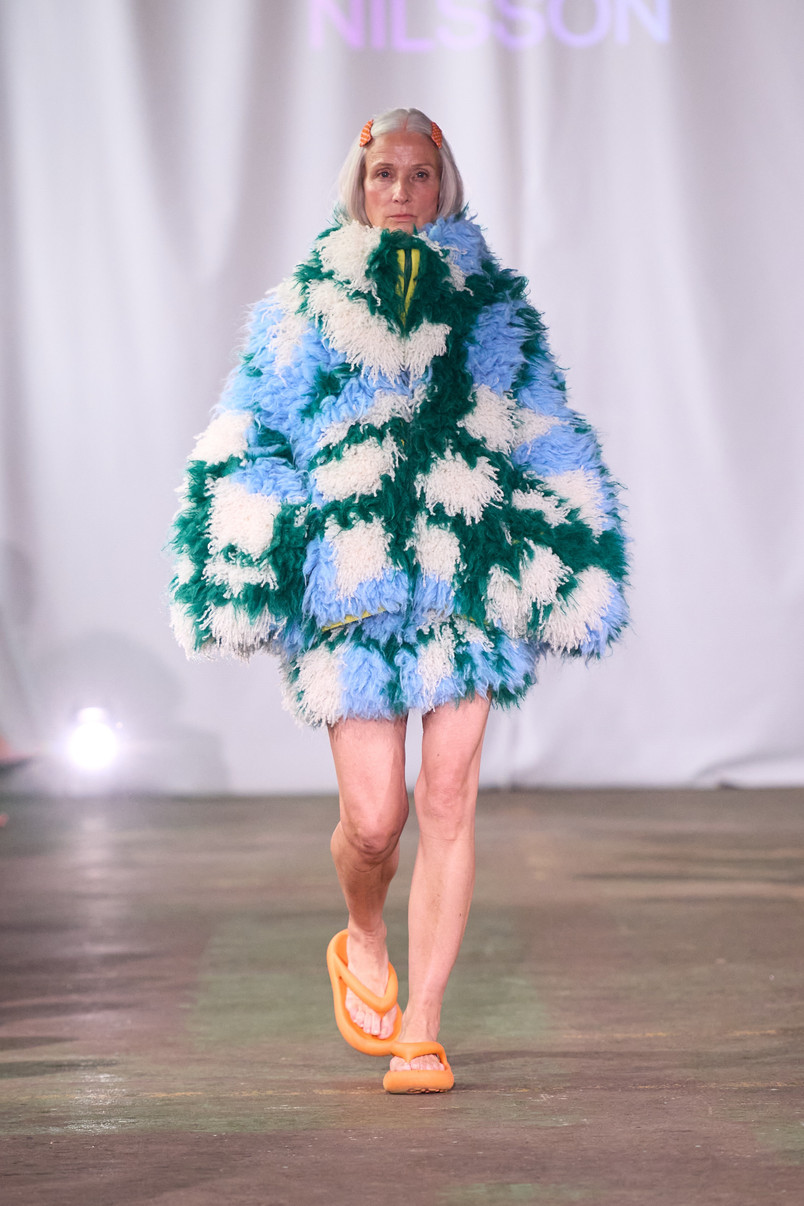 Model på catwalk iført en voluminøs, langhåret jakke i lyseblå, grønne og hvide nuancer samt orange sandaler.