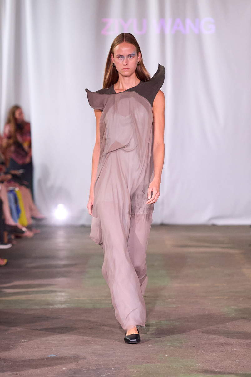 Model på catwalk iført en draperet grå kjole med skulpturelle sorte skulderdetaljer.
