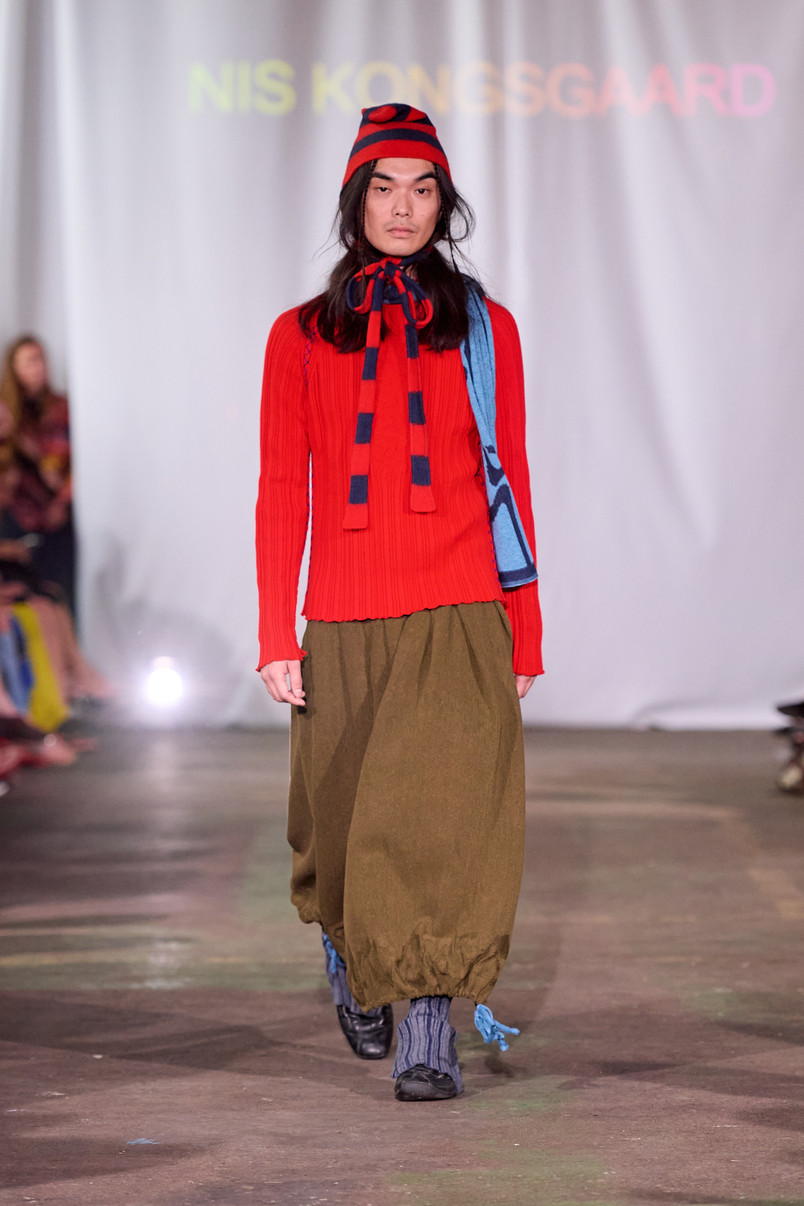 Model på catwalk iført rød ribstrikket trøje, rød- og blåstribet hue og tørklæde samt olivengrønne ballonbukser.