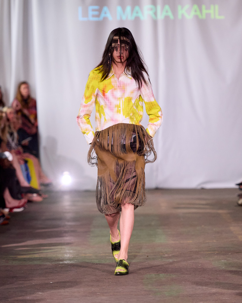 Model på catwalk iført gul- og lyserødmønstret skjorte og frynset nederdel med ansigtsdækkende perlehovedsmykke.