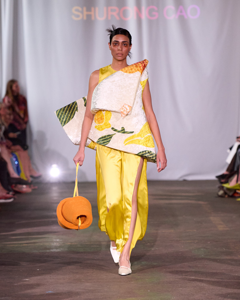 Model på catwalk iført skulpturel top med citrusmønster, gule bukser og en rund orange taske.