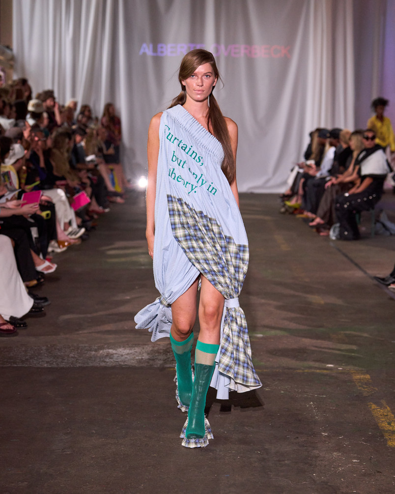 Model på catwalk iført lyseblå draperet kjole med teksttryk, ternede detaljer og turkise knæstrømper.