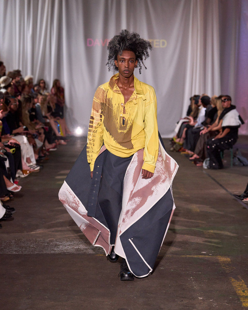Model på catwalk iført gul mønstret skjorte og voluminøs nederdel med grafiske paneler.