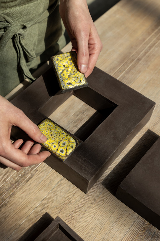 hands holding two yellow glaze tests over a unglazed clay object