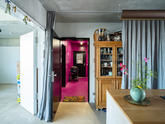 ndgang til en lejlighed med en åben dør, der fører til en gang med pink vægge og gulv. Til højre står et vitrineskab og en bordplade med en vase med blomster